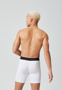 Boxer aderenti bianchi con elastico nero in vita, design attillato, texture liscia e lunghezza fino alla metà della coscia, mostrati su un modello maschile visto da dietro.