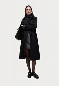 Svart trenchcoat med dubbelradiga knappar och midjebälte, bärs över en grå kjol. Modellen håller en svart handväska och har på sig svarta skor och strumpbyxor.