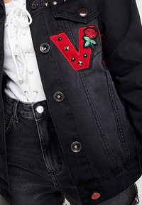 Czarna kurtka jeansowa z czerwoną, wyszywaną literą "V" i różą, srebrnymi ćwiekami, zapinana na guziki, o fakturowanej powierzchni. Noszona na białej bluzce i czarnych dżinsach.