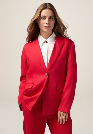 Blazer rosso con due tasche frontali, chiusura a un bottone e rever strutturati. Indossato sopra una camicia bianca con un dettaglio dorato. Texture liscia.