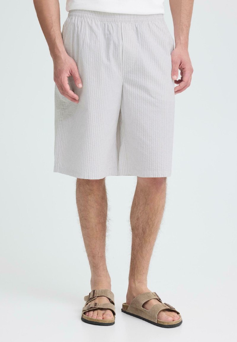 Homme portant un short rayé gris clair atteignant les genoux et des sandales en suède beige, debout devant un fond blanc uni.