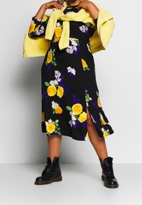 Robe noire à fleurs avec des roses jaunes, des accents violets et blancs, dotée d'une fente sur le côté. Associée à des bottes montantes noires à lacets et un pull jaune noué à la taille.