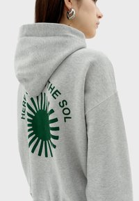 Grijze hoodie met een groene zonafbeelding op de achterkant en de tekst "HERE FOR THE SOL." Zachte textuur en een ontspannen pasvorm.