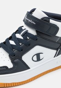 High-top sneaker with navy leather and white textile, featuring a velcro strap, navy laces, and a textured white panel. Rubber sole with orange accents.