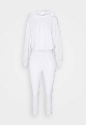 Biały dwuczęściowy strój damski składający się z krótkiej bluzy z kapturem i wysokim stanem spodni dresowych ze sznurkiem w pasie.