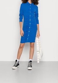 Sininen, pitkähihaiset paitamekko, jossa on kauluksellinen pääntie ja napitettu etuosa. Mekossa on sivuilta poimittu yksityiskohta, yhdistetty mustien korkeavartisten tennareiden kanssa.