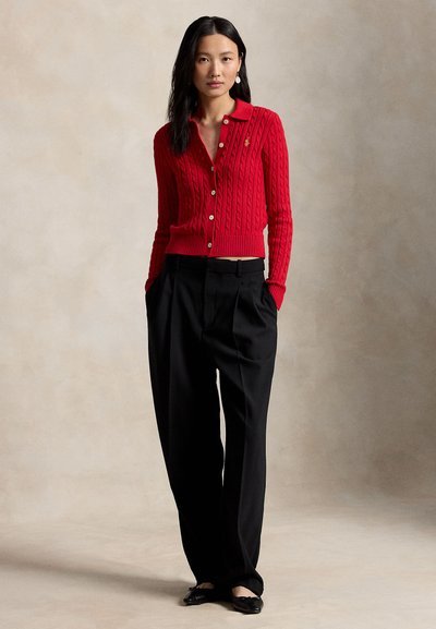 Kobieta ubrana w czerwony, zapinany na guziki, sweter warkoczowy, czarne spodnie o szerokich nogawkach oraz czarne baleriny, stojąca na tle jednolitego beżowego tła.