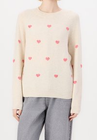 Sweter w kolorze kremowym, wykonany z miękkiego materiału, z różowymi wzorami serc rozmieszczonymi równomiernie na przodzie. Okrągły dekolt i ściągacz na dole.