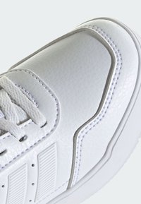 Zapatilla blanca con parte superior sintética texturizada, diseño curvo, costuras en contraste y un panel acentuado acanalado en el lateral.