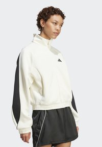 White zip-up jacket with black accents on sleeves, featuring a fitted hem and a small logo. Worn with black shorts, showcasing sporty design.