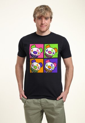 Jonge man die een zwart T-shirt draagt met vier kleurrijke clowngezichten in een raster, elk met verschillende achtergrond- en maskerkleuren.