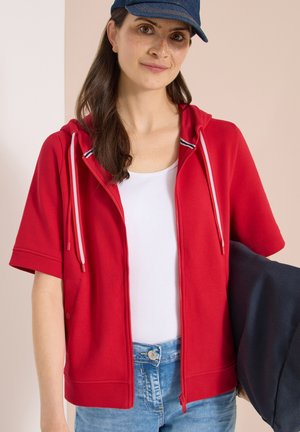 Młoda kobieta w niebieskiej czapce, czerwonej bluzie z zamkiem i krótkim rękawem, białej koszuli i dżinsach, trzymająca ciemną kurtkę na ramieniu.