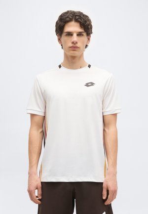 Fiatal férfi hullámos sötét hajjal, fehér sportpólóban, színes csíkokkal és geometriai logóval a mellkasán.