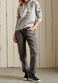 Felpa grigia con logo bianco sul davanti, abbinata a jeans grigio scuro. Indossata con sneaker alte nere. Sullo sfondo, pannellatura in legno chiaro.