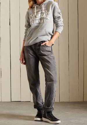 Felpa grigia con logo bianco sul davanti, abbinata a jeans grigio scuro. Indossata con sneaker alte nere. Sullo sfondo, pannellatura in legno chiaro.