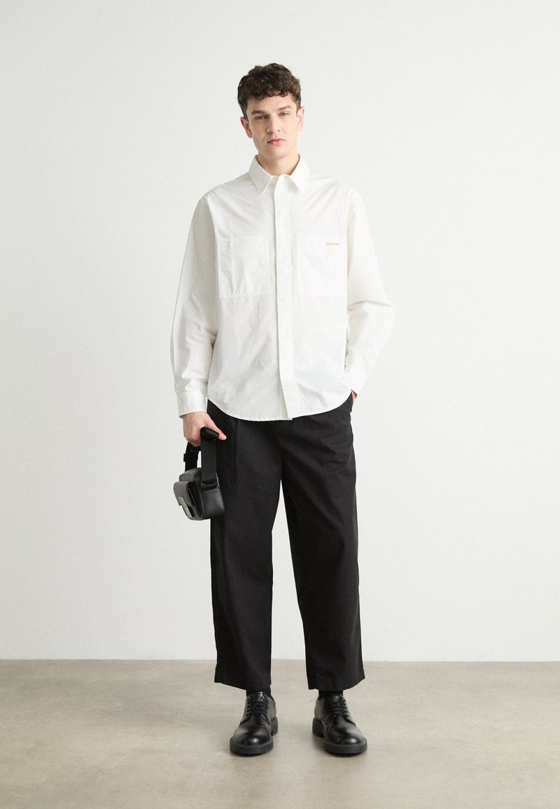 Camicia bianca a maniche lunghe con due tasche sul petto e logo dorato, abbinata a pantaloni neri larghi e scarpe nere con lacci, mentre tiene una piccola borsa.