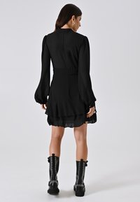 Schwarzes Kleid mit langen Ärmeln, Rüschensaum und taillierter Taille. Tragen mit kniehohen schwarzen Stiefeln mit Nieten-Details. Glatte Stoffoberfläche.
