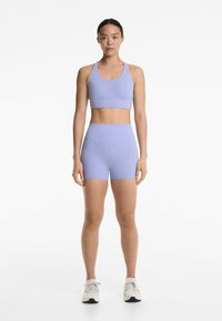Set de sutien sport și pantaloni scurți de culoare purpurie deschisă, dintr-un material elastic. Design simplu, cu o siluetă ajustată și fără modele vizibile. Se poartă pantofi sport.