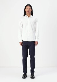 Camisa blanca de manga larga con cierre de botones y un cuello puntiagudo, combinada con pantalones azul marino y zapatos negros, de pie frente a un fondo simple.