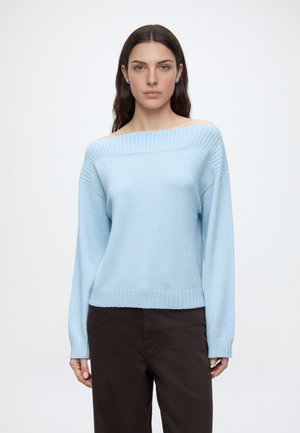 Femme aux longs cheveux foncés portant un pull bleu clair épaules dénudées et un pantalon marron foncé, debout devant un fond blanc uni.