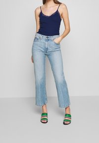 Kvinna iförd en marinblå spetskantad linne, ljusblå raka croppade jeans och gröna randiga öppna klackar, stående med en hand i fickan.