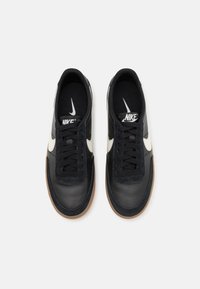 Ett par svarta Nike-sneakers med vita swoosh-loggor, svarta snören och sålar i gumfärg, sedd uppifrån mot en vit bakgrund.