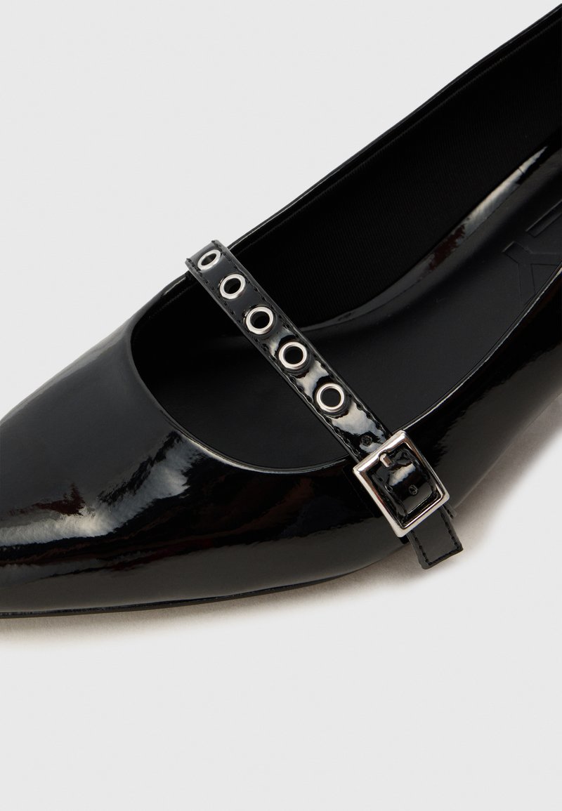 Zapato Mary Jane de charol negro con punta afilada, que presenta una tira con hebilla y detalles de cortes circulares a lo largo de la tira.