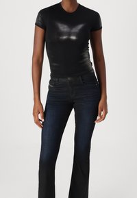 Nainen, jolla on päällään vartalonmyötäinen lyhythihainen musta paita ja tummansiniset korkeavyötäröiset levenevät farkut, seisoo yksinkertaista taustaa vasten.