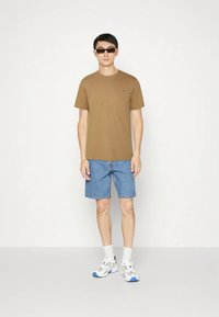 Ruda trumpomis rankovėmis marškinėliai su mažu logotipu, derinami su džinsiniais šortais ir stambiais baltais sportbačiais su mėlynomis detalėmis.