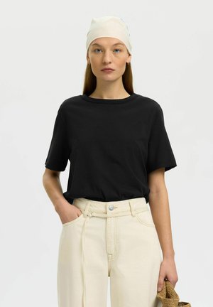 Woman with light skin and blue eyes wears a white headscarf, black short-sleeve shirt, and high-waisted cream pants, holding a woven item.