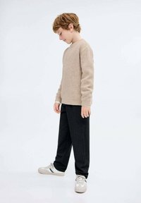 Svetlo béžový textúrovaný pletený sveter, čierne zúžené nohavice a šedé tenisky s modrými pruhmi; dieťa stojí v bočnom pohľade a pozerá sa dolu.