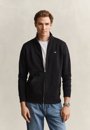 Jeune homme portant une veste noire zippée par-dessus une chemise blanche et un jean bleu, debout devant un fond clair uni.