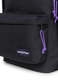 Mochila de tela negra con un detalle en púrpura. Cuenta con un bolsillo frontal, cierre con cordón y una etiqueta con logo en la parte inferior.