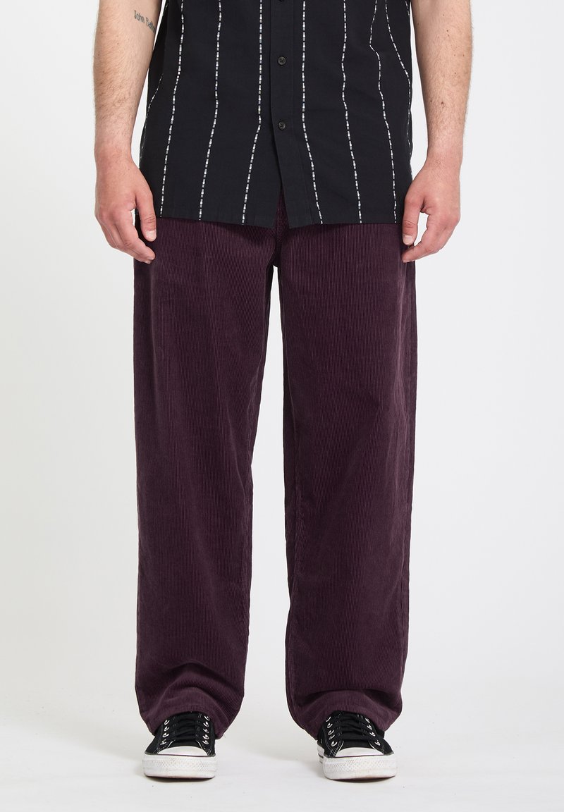 Homme portant un pantalon en velours côtelé violet foncé ample, des baskets noires et blanches, et une chemise noire à boutons avec des rayures blanches verticales.