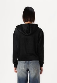 Sudadera negra con capucha, de textura suave y corte relajado, combinada con vaqueros azules desgastados que presentan un característico cosido en los bolsillos traseros.