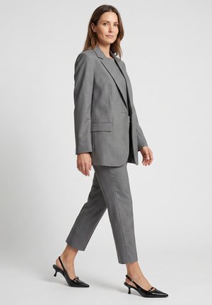 Kvinde iført gråt skræddersyet jakkesæt med korte bukser og sorte slingback sko med lav kittenhæl mod ensfarvet baggrund.