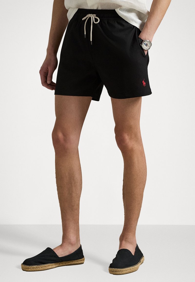Svart badeshorts med elastisk midje og snor, med en liten rød logo på venstre side. Glatt stoff og avslappet passform.