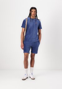 Chemise à manches courtes en velours côtelé bleu et shorts assortis avec une texture côtelée, associée à des baskets blanches et des chaussettes blanches, ainsi qu'un sac à bandoulière beige.