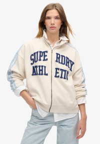 Sudadera cremosa con cremallera, con letras bordadas en azul "SUPERDRY ATHLETIC" y rayas azul claro en las mangas. Presenta puños y dobladillos acanalados.