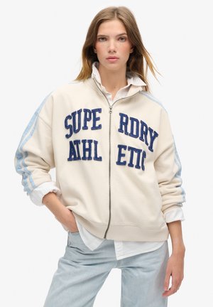 Crème sweatvest met ritssluiting en blauwe geborduurde lettering "SUPERDRY ATHLETIC" en lichtblauwe strepen op de mouwen. Heeft ribbels aan de manchetten en onderkanten.