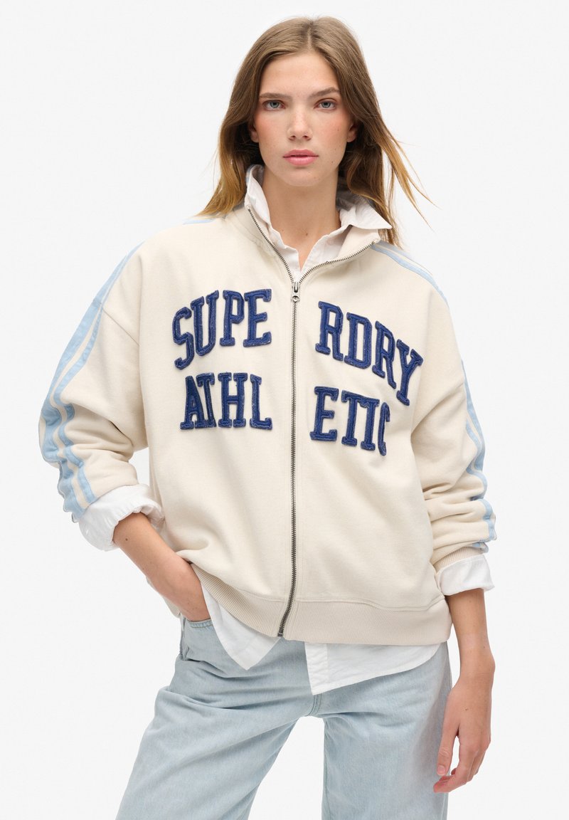 Sudadera cremosa con cremallera, con letras bordadas en azul "SUPERDRY ATHLETIC" y rayas azul claro en las mangas. Presenta puños y dobladillos acanalados.