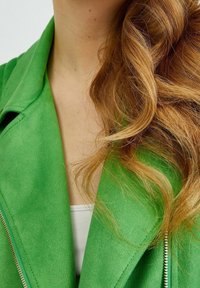 Donna con lunghi capelli ondulati rossi che indossa una giacca verde brillante sopra una maglietta bianca, vista dalla nuca al petto.