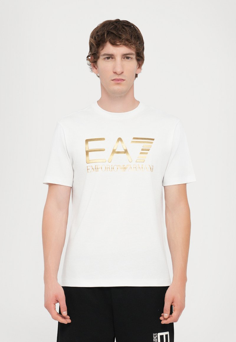 Hvit bomulls T-skjorte med en skinnende gull logo "EA7 EMPORIO ARMANI" over brystet, rund halslinning og korte ermer.