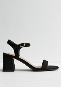 Svart klackad sandal med bred klack, sammetsliknande textur, ankelrem med metallspänne och platt framrem.