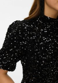 Melnas pērļu topa ar augstu apkakli, īsām pūkainām piedurknēm un strukturētu audumu. Iezīmēts ar daudzām spožām, apaļām pērļu, sniedzot atstarojošu efektu.