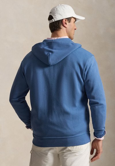 Mees, kes kannab sinist kapuutsiga pusa, valget nokamütsi ja heledaid pükse, selg vastu neutraalset tausta.