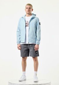 Light blue lightweight jacket with hood, gray shorts, and white sneakers. Features elastic cuffs, front zip, and graphic print on white t-shirt.