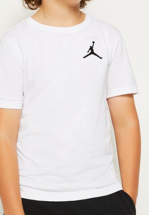 Child wearing a white t-shirt with a black Jumpman logo on the left chest, standing with hands in pockets.