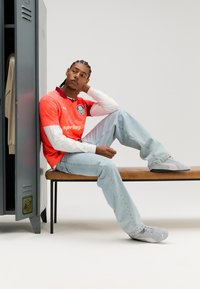 Rdeč in bel dolg rokav dresa z logotipom, modre jeans hlače in sive superge. Sedi na leseni klopi poleg omarice.