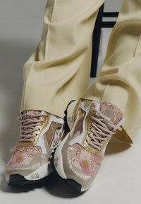 Pantalones crema y zapatillas de deporte lace-up en dorado-rosa con parches decorativos y paneles texturizados sobre un fondo claro.